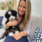 Woman sitting on a couch, smiling, and holding a small black and white puppy on her lap.